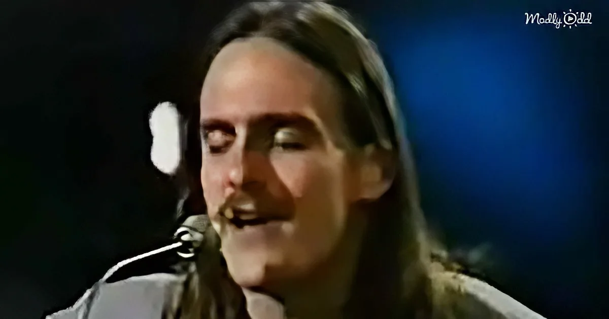 A person with long hair and a beard sings into a microphone, wearing a light-colored shirt.