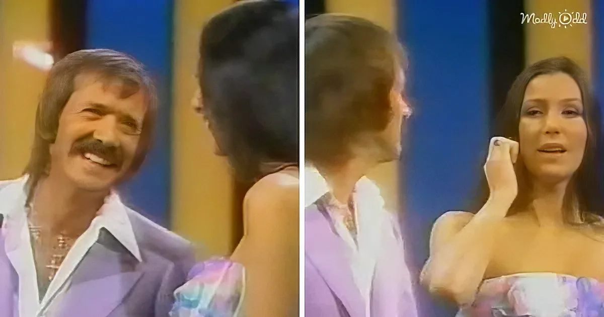 A man in a purple suit smiles at a woman in a strapless dress, who is holding a tissue to her face.