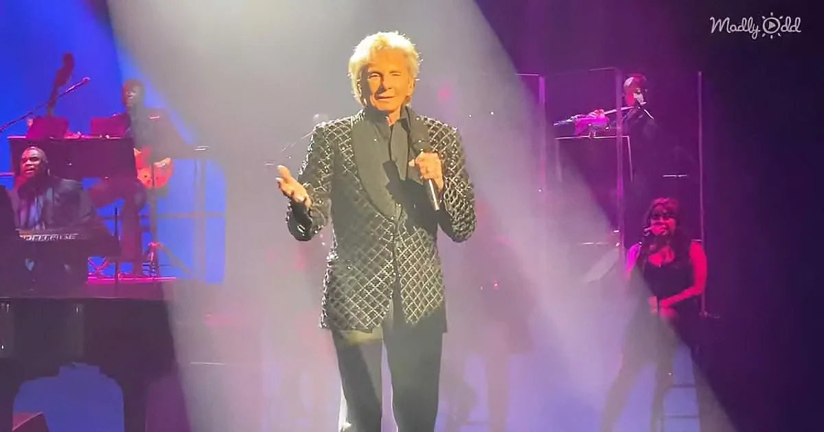 A man in a black patterned suit sings on stage with a band and microphone.