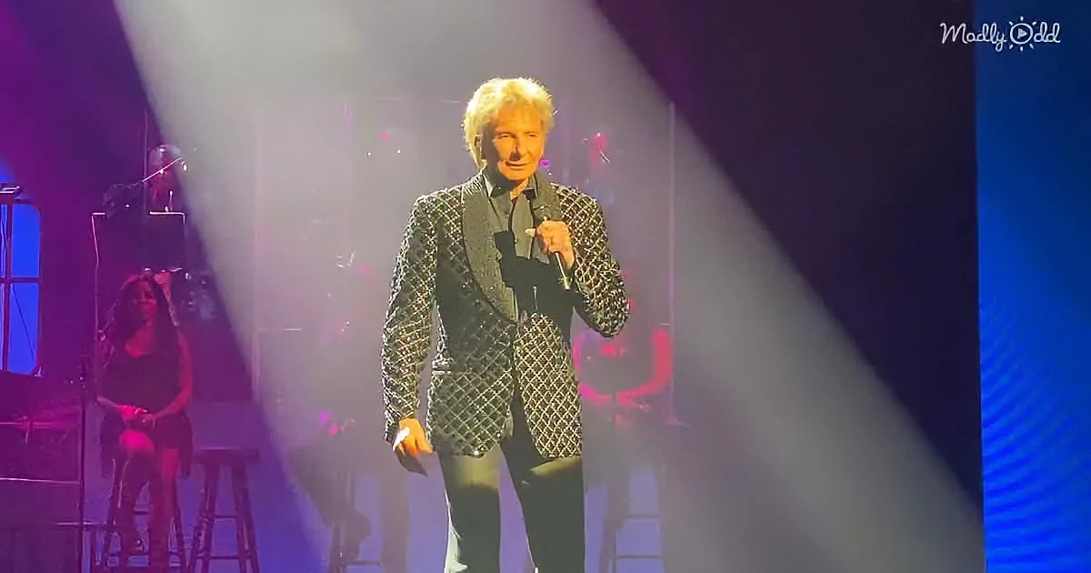 A man in a patterned suit holds a microphone on stage, with colorful lighting and a seated figure in the background.