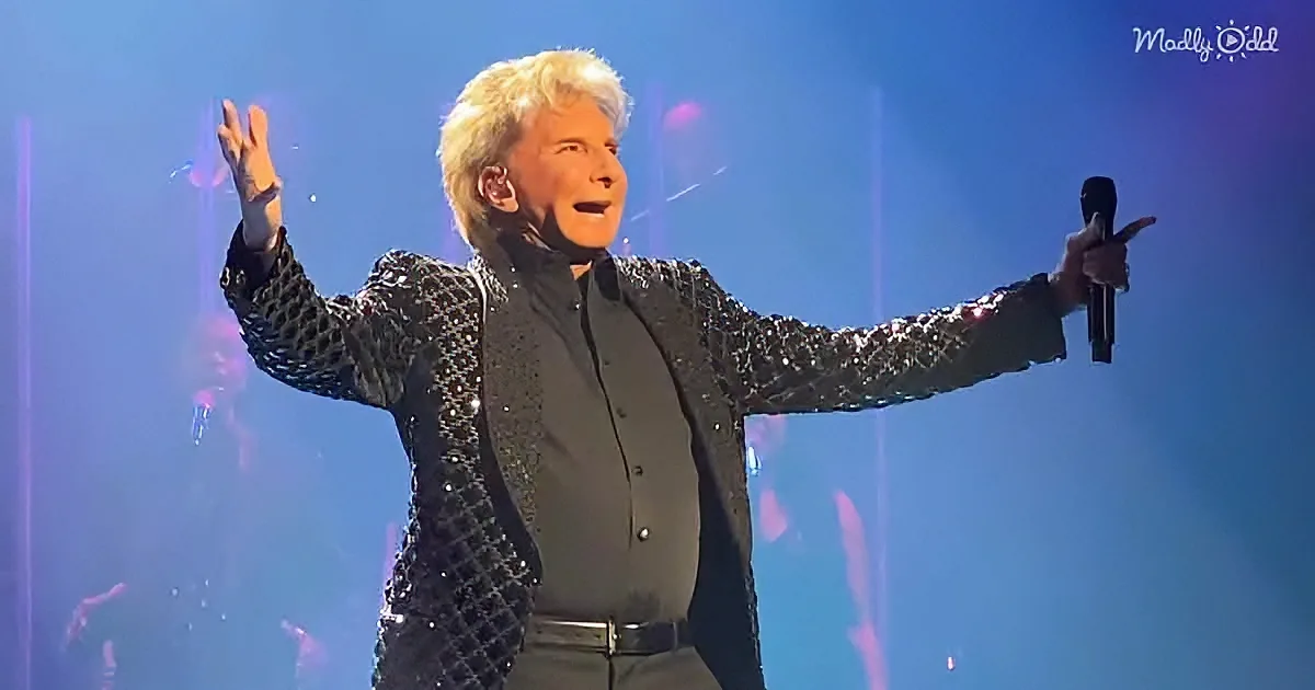A performer in a glittery black outfit sings passionately on stage with arms outstretched.