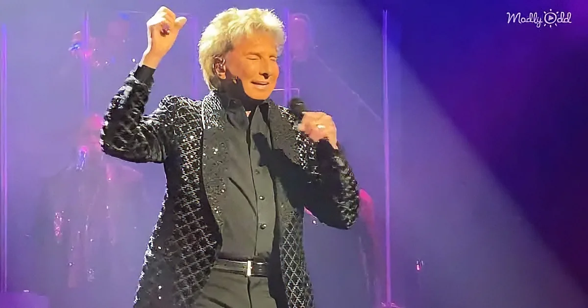 A performer in a glittery jacket sings into a microphone on stage with purple lighting.