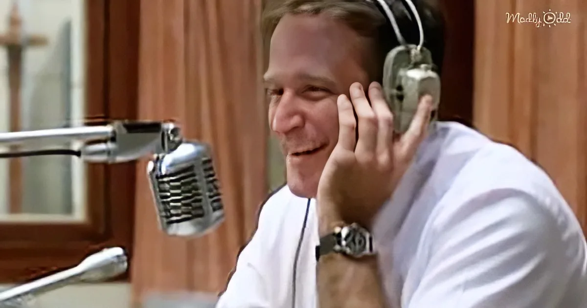 A man in a recording studio wearing headphones, smiling, with a microphone in front of him.