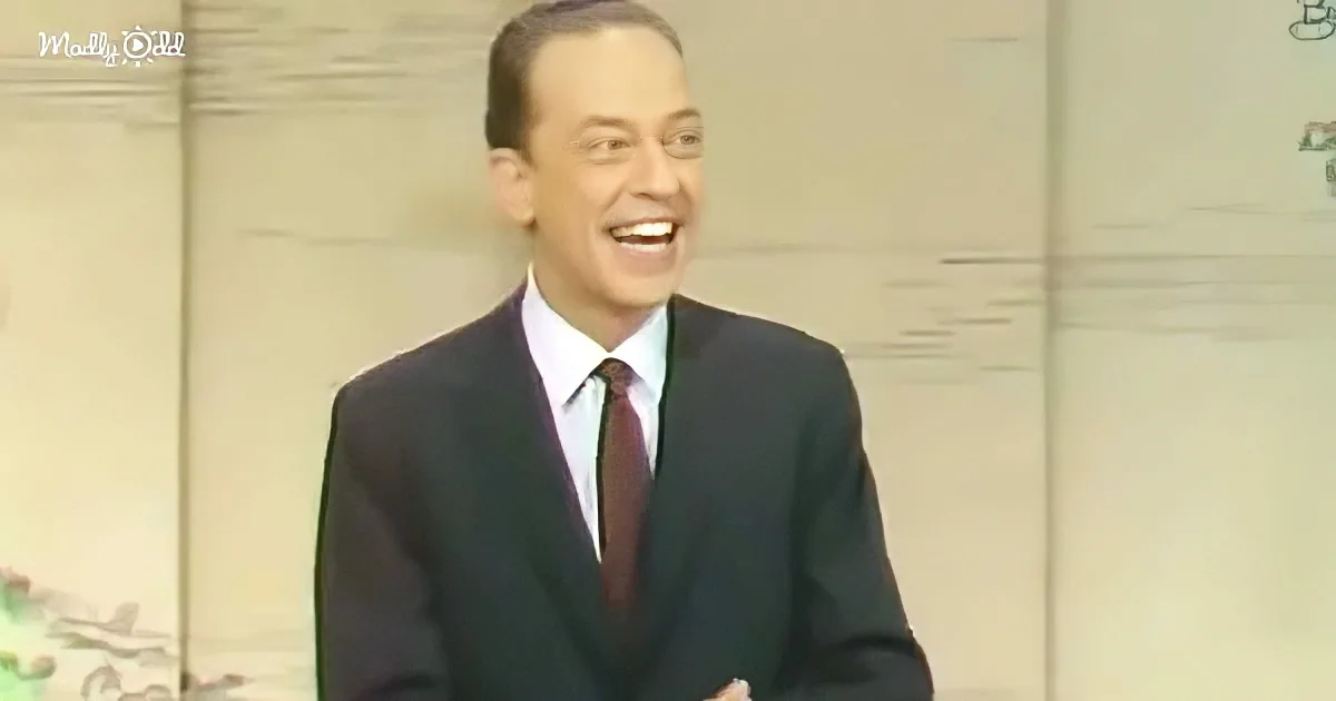 A man in a dark suit and tie is smiling widely, standing against a light background.
