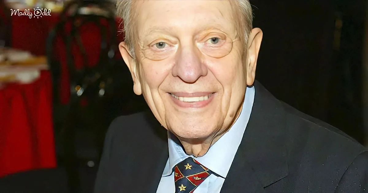 An older man in a suit and tie smiles warmly, wearing a medal, against a red-clothed table backdrop.