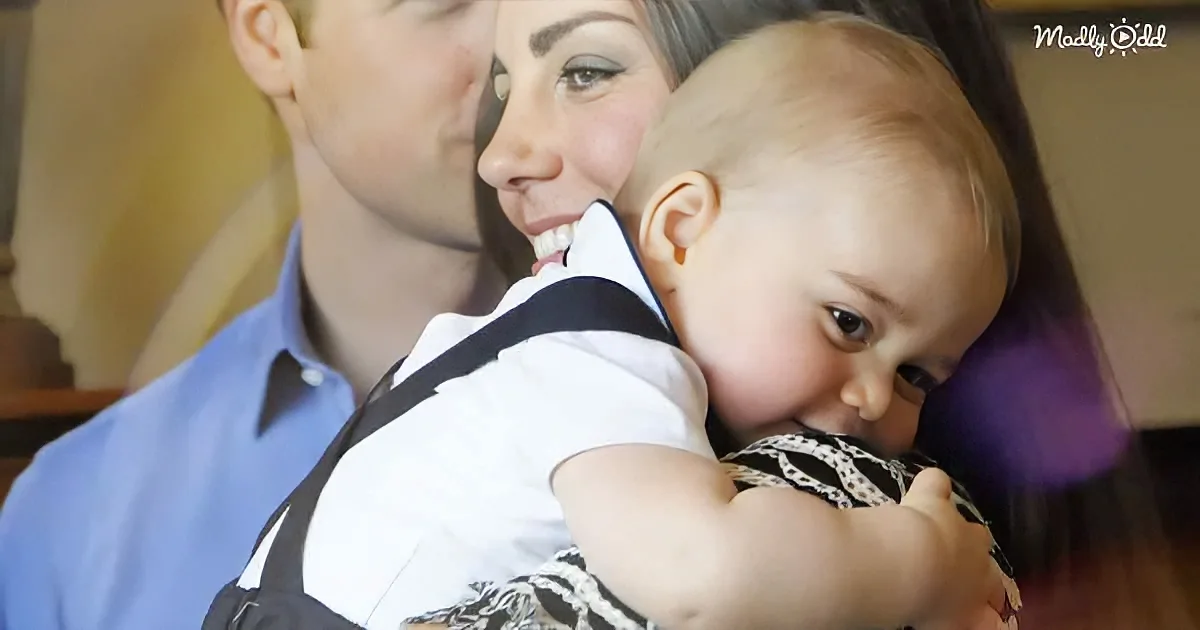 A woman holds a baby in a carrier, smiling, with a man in a blue shirt nearby.