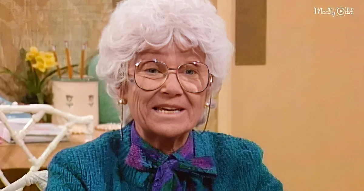 An elderly woman with white hair and glasses smiles, wearing a blue sweater and purple shirt, in a cozy setting.