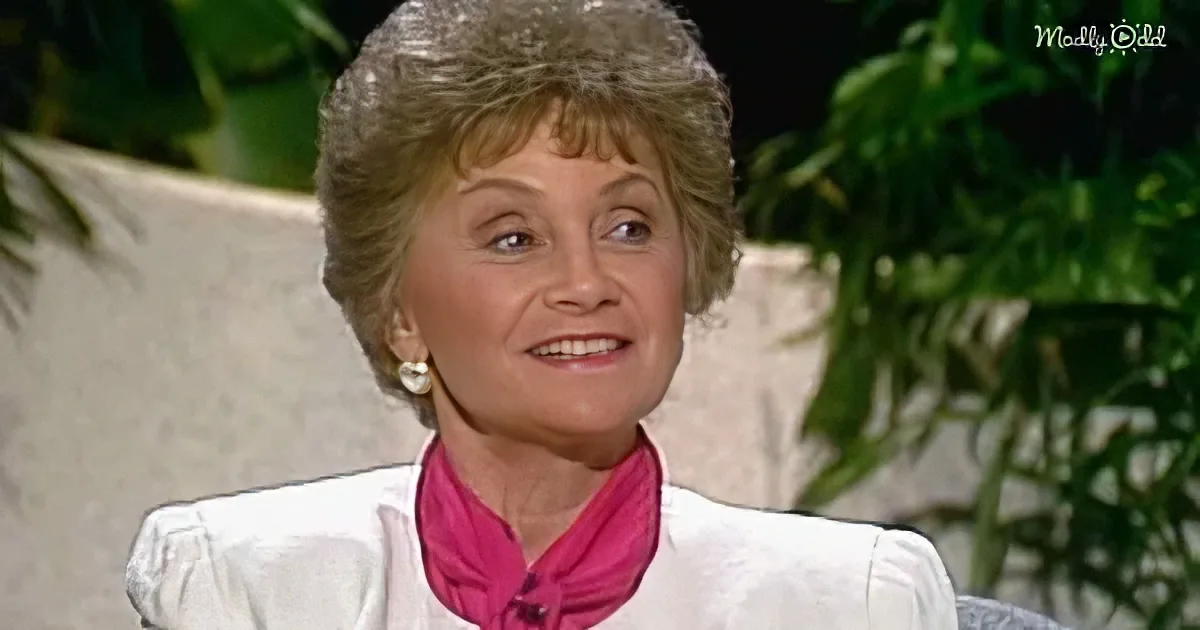 A woman with short, styled hair, wearing a white blouse and a pink scarf, smiles while seated in front of greenery.