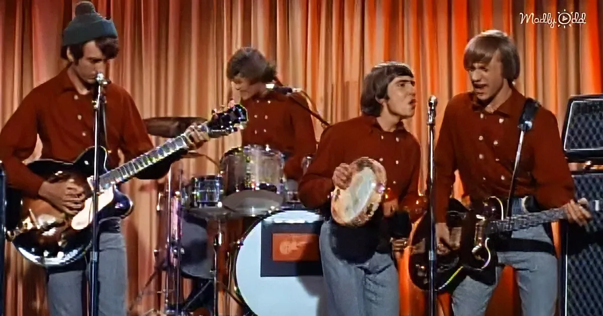 Four musicians in brown shirts perform on stage, each with their instrument, against a backdrop of orange curtains.