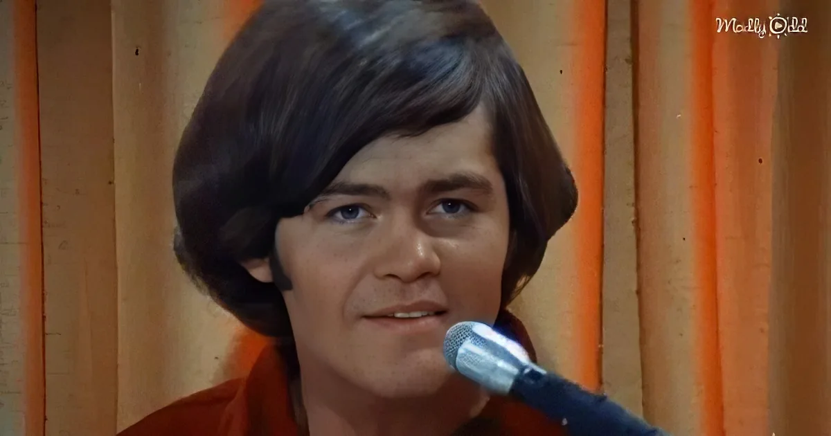 A person with shoulder-length hair, wearing a red shirt, is speaking into a microphone in front of a wooden background.