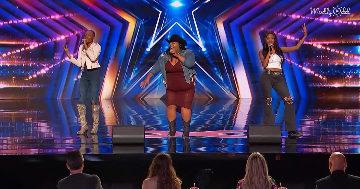 Three women perform on stage with microphones, wearing stylish outfits, under vibrant lighting.