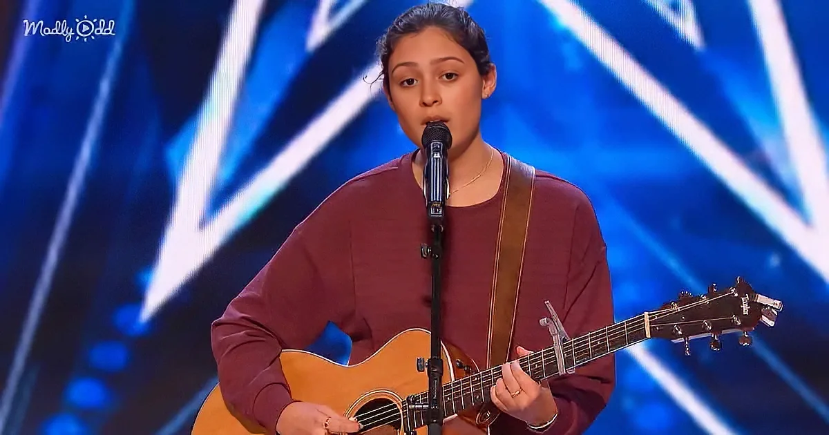 A person in a maroon shirt plays an acoustic guitar on stage with a microphone.
