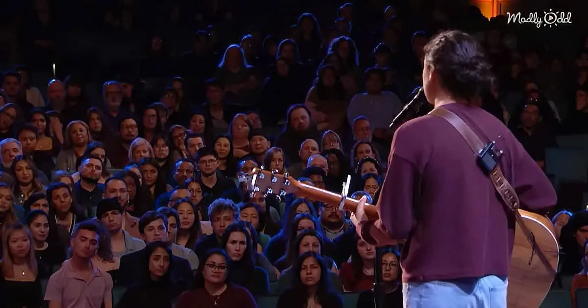 A person in a purple shirt plays guitar on stage in front of a large audience.