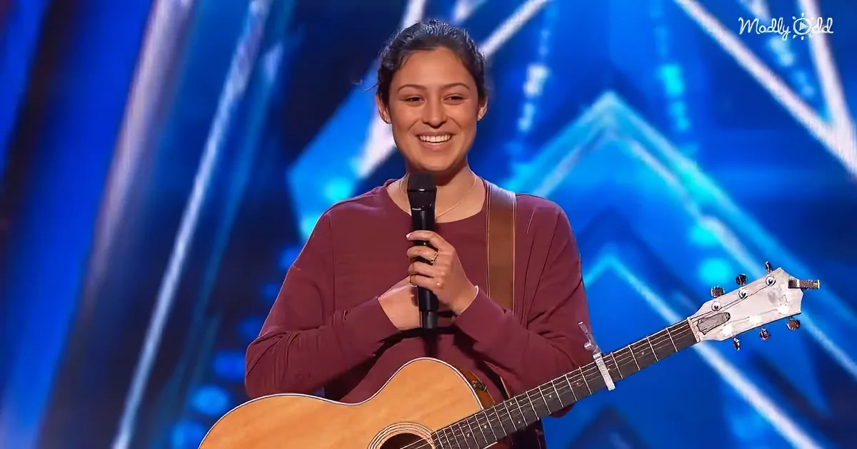 A woman smiles on stage, holding a microphone and an acoustic guitar, against a blue geometric backdrop.