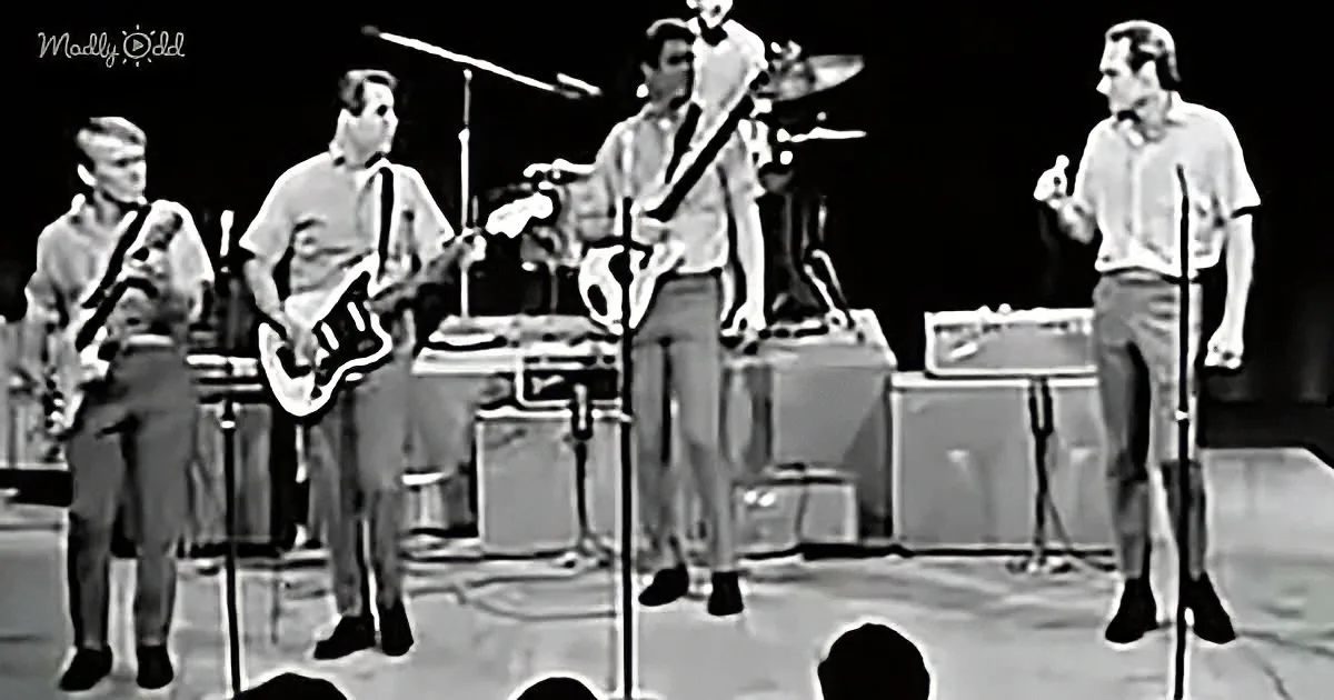 A band performs on stage with guitars, drums, and a keyboard, all in a black-and-white setting.