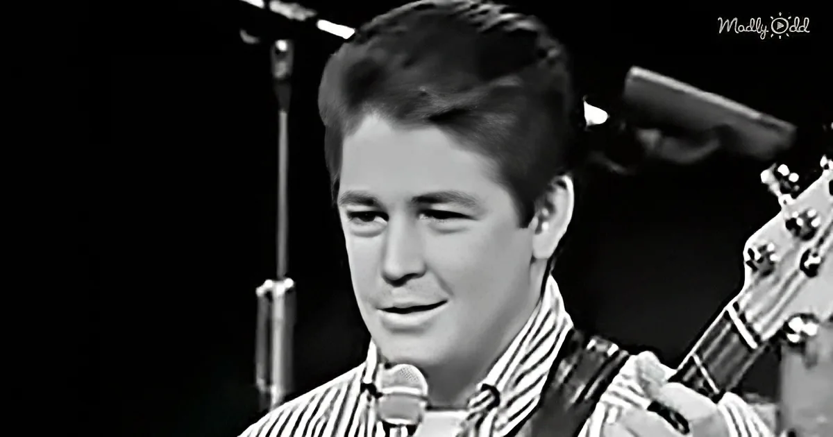 A man in a striped shirt plays guitar and sings into a microphone.