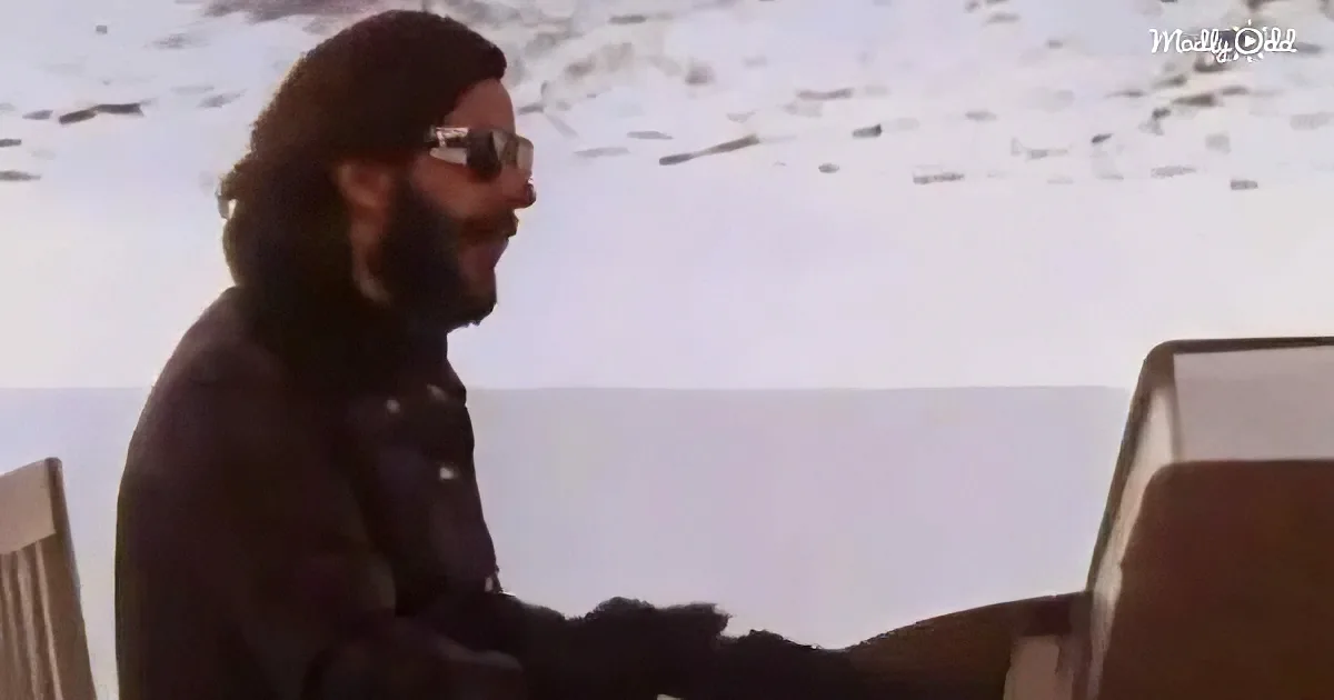 A man with long hair and beard, wearing sunglasses and a dark jacket, sits at a piano, playing.