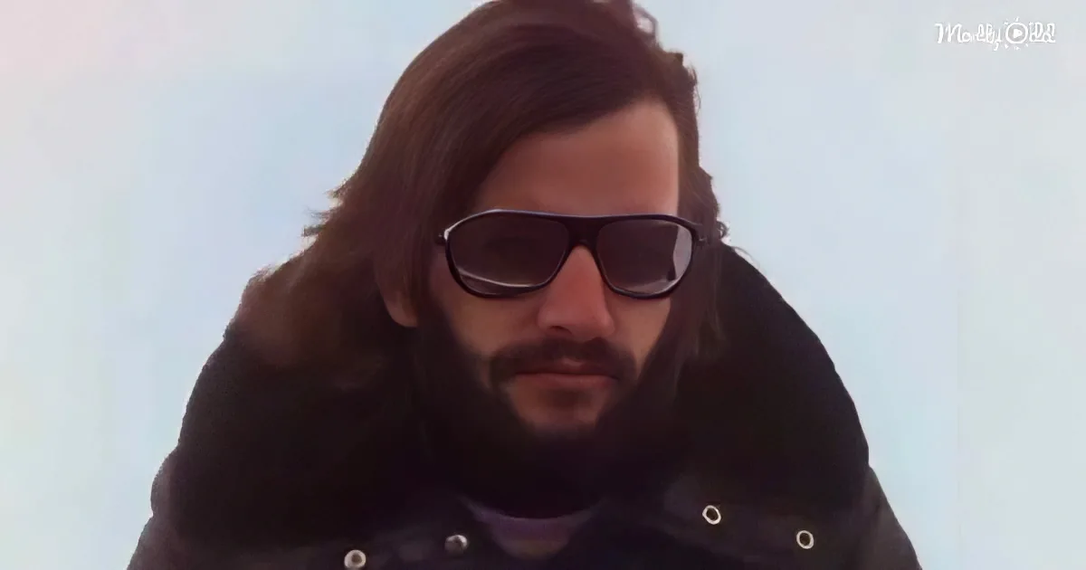 A man with long hair and a beard wears sunglasses and a dark jacket against a light sky.