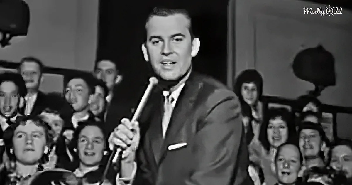 A man in a suit holds a microphone, surrounded by a cheering crowd in a black-and-white setting.