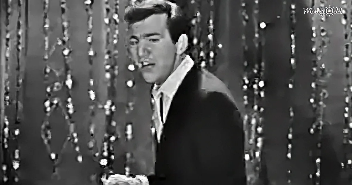 A man in a suit stands in front of a shimmering backdrop, holding a microphone.