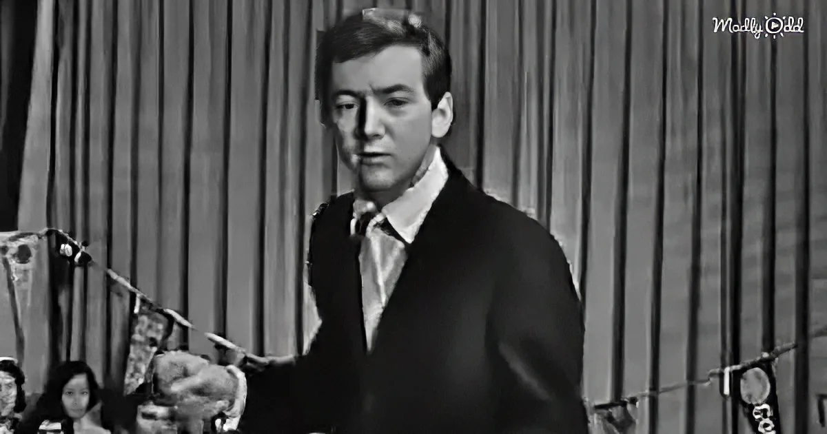 A man in a suit and tie stands in front of microphones, gesturing with his right hand.