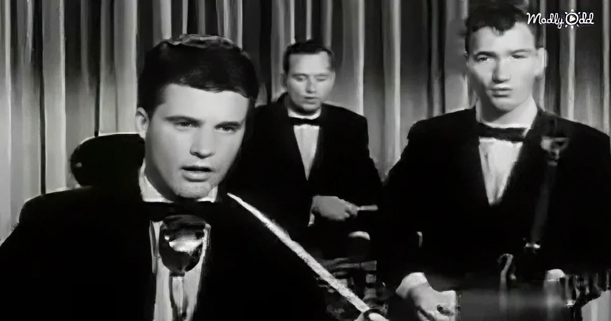 A man in a tuxedo sings into a microphone, accompanied by two other men in formal attire.