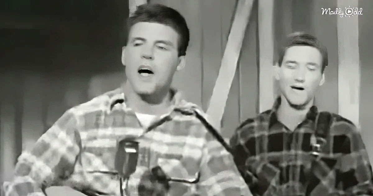 Two men in plaid shirts sing into microphones, one in the foreground, the other slightly behind.