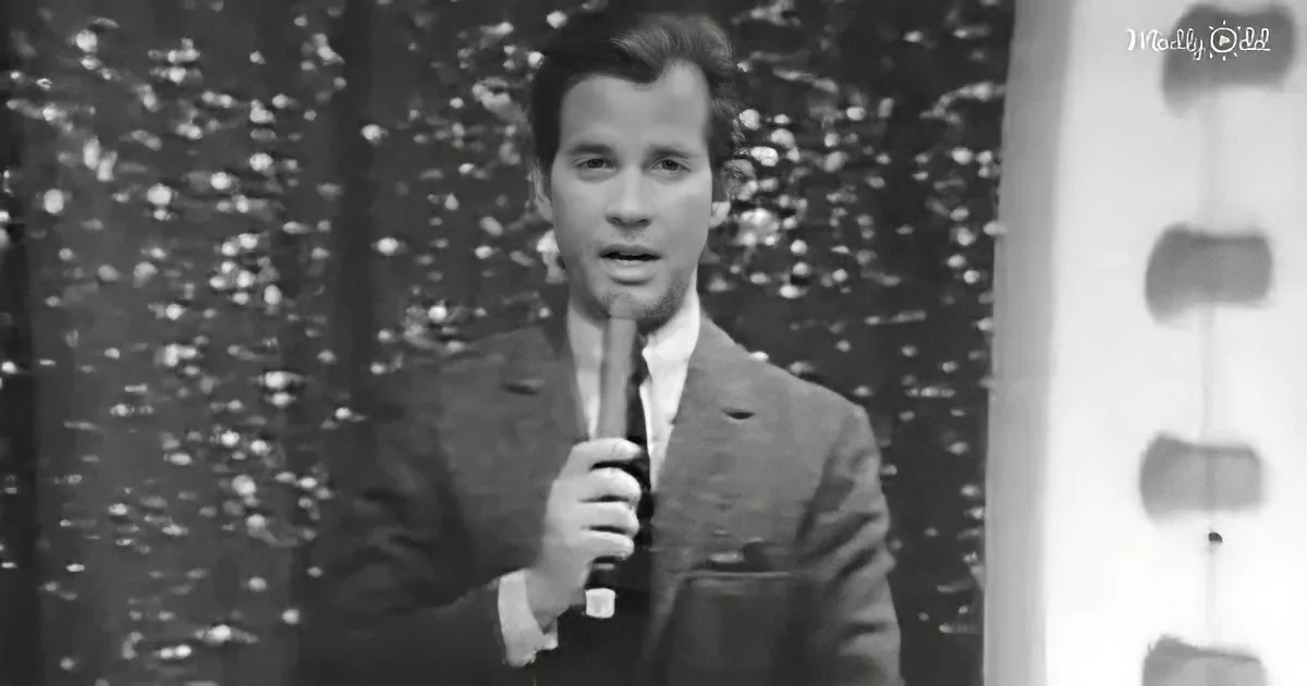 A man in a suit holds a microphone, standing in front of a patterned backdrop.