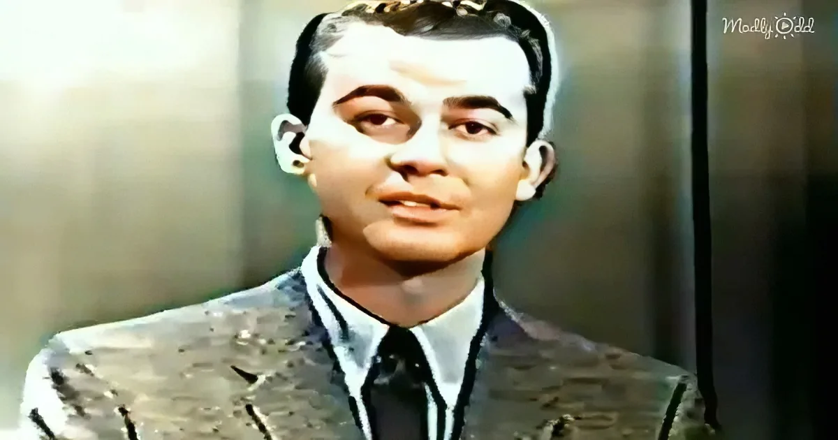 A man in a suit and tie, with a serious expression, stands against a blurred background.