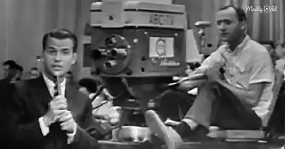 A man in a suit holds a microphone, another adjusts a large camera in the background.