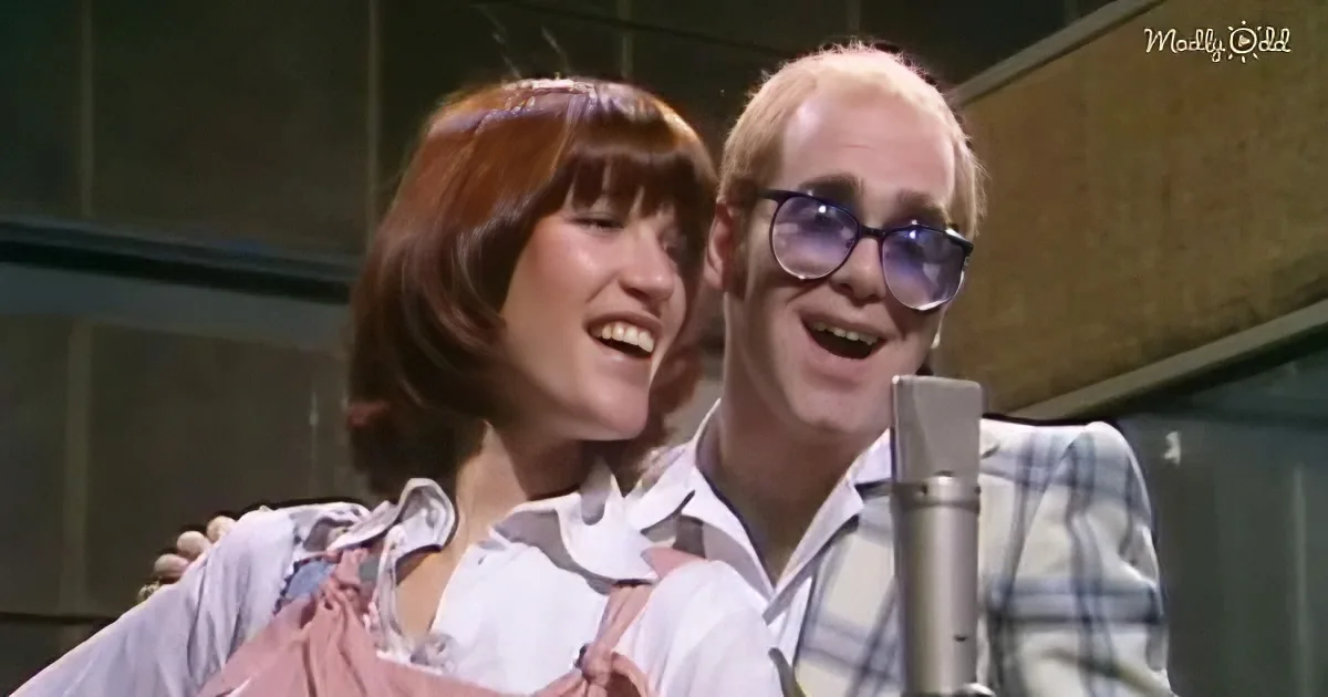 Elton John and a woman in a pink dress share a joyful moment, both smiling near a microphone.