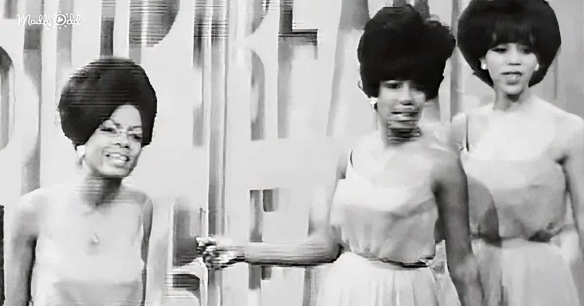 Three women in 1960s attire, one holding a microphone, standing in front of a backdrop.