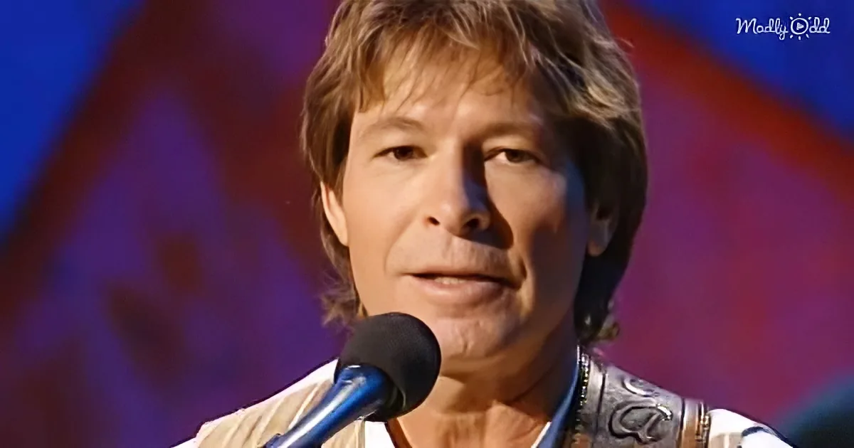 A man with shoulder-length hair speaks into a microphone, wearing a patterned shirt, against a blurred background.