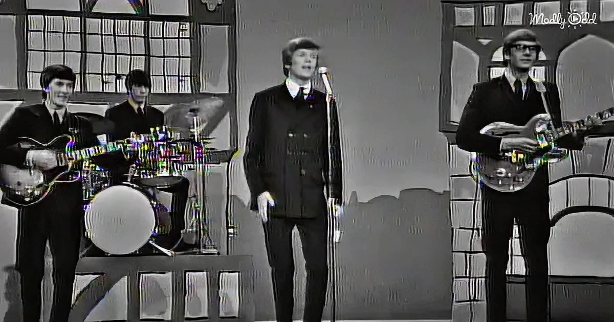 A man in a suit sings into a microphone, accompanied by a band playing guitars and drums.