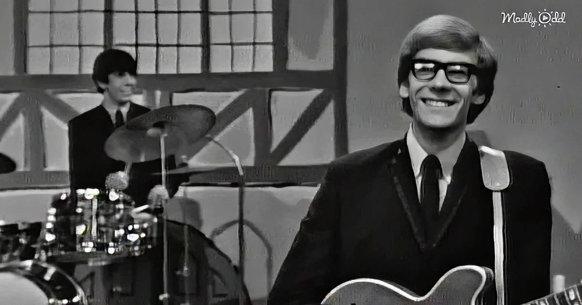 A smiling man in a suit holds a guitar, with a drummer in the background, both in a studio setting.