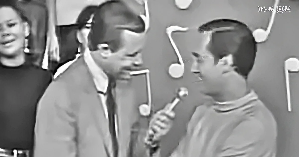A man in a suit holds a microphone, speaking to another man in a casual setting.
