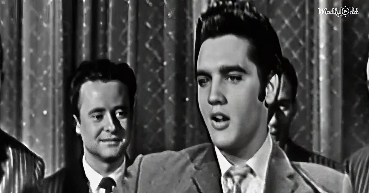Elvis Presley in a suit and tie, singing passionately, with another man in the background.