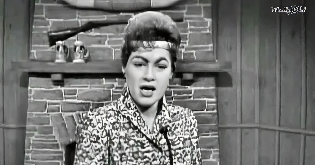 A woman in a floral shirt stands in front of a brick wall, mouth open, seemingly speaking or singing.
