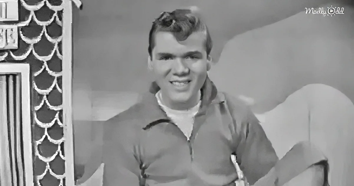 A man in a polo shirt smiles, sitting on a couch with a decorative wall behind him.