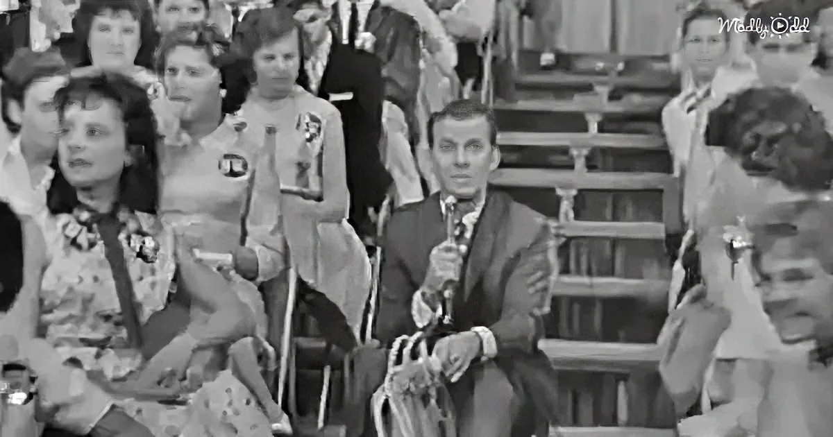 A man in a suit holds a microphone, seated in front of a crowd at a formal event.