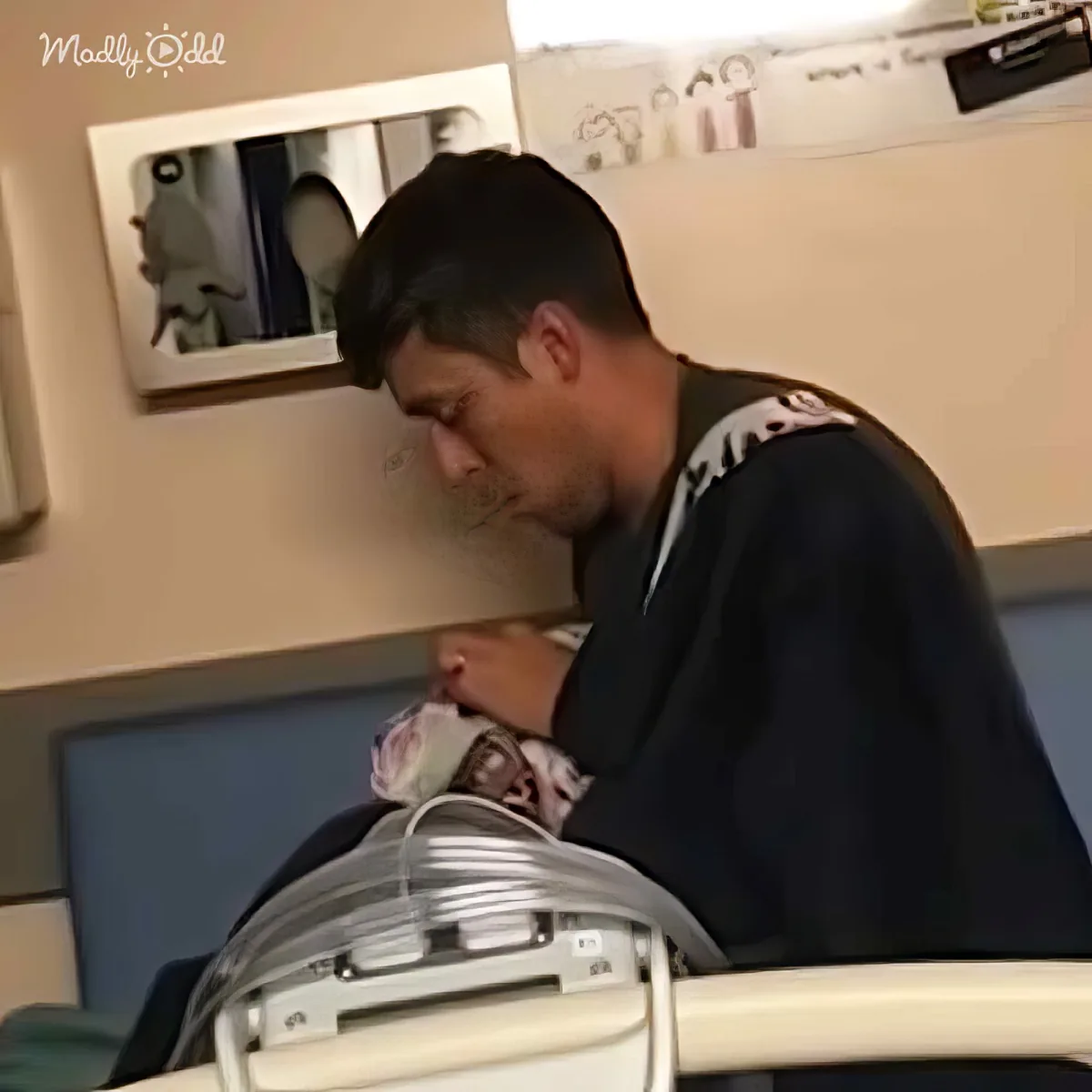 A man leans over a baby in a hospital bassinet, holding the baby's hand.