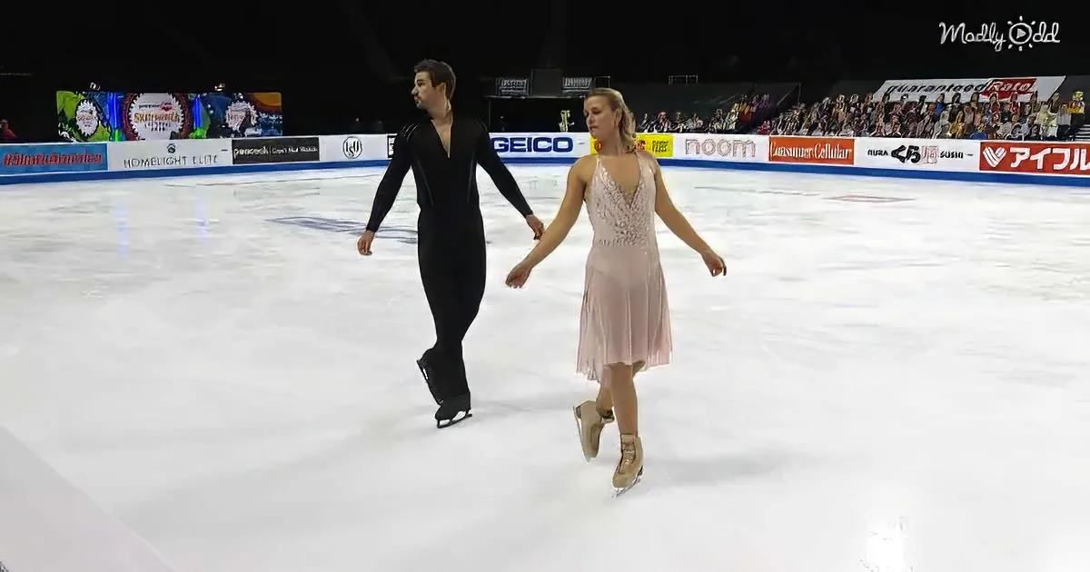A figure skating couple glides on ice, holding hands, with a crowd and sponsor banners in the background.