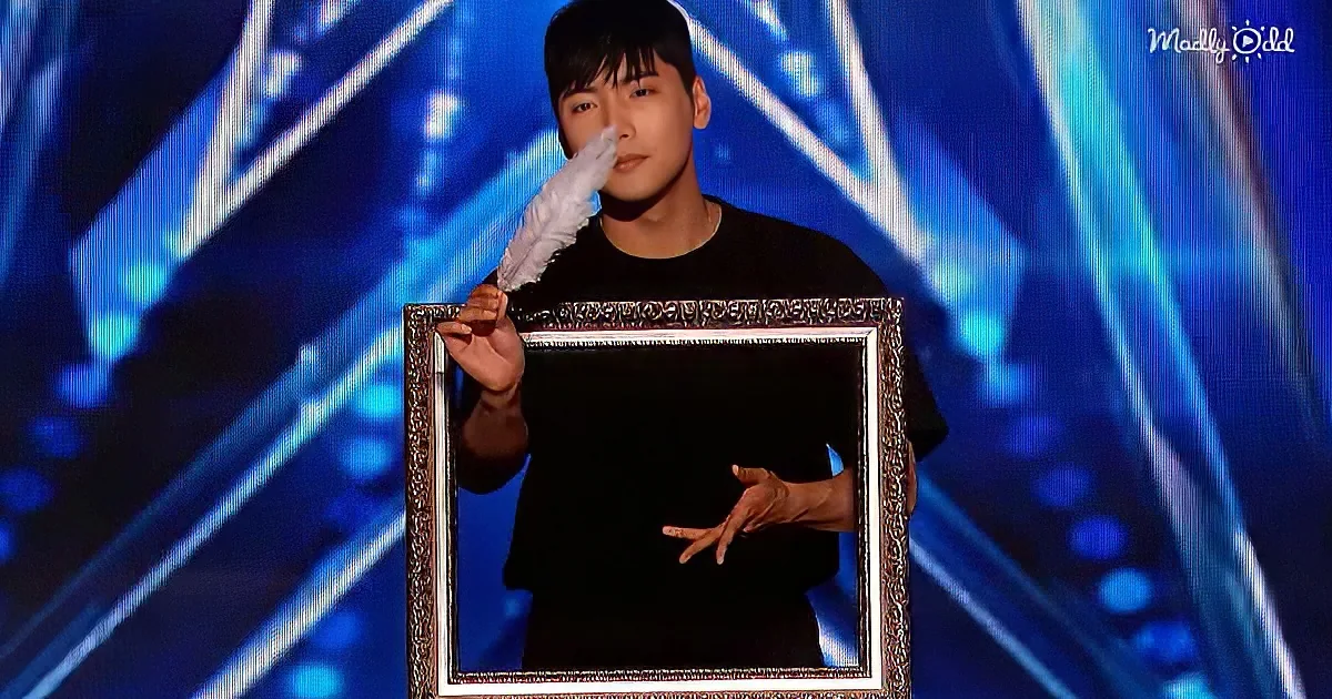 A person in a black shirt holds a feather duster and a large empty frame on a stage with blue lights.