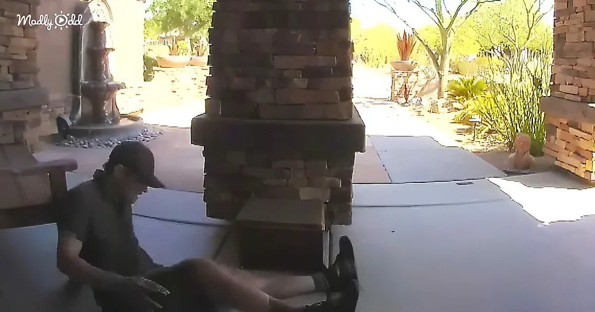 A man in a cap sits on a patio, looking down, with a brick pillar and outdoor decor in the background.