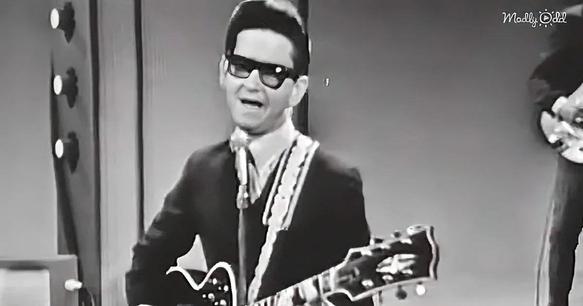 A man in a suit and sunglasses plays a guitar, wearing a white scarf, in a black-and-white image.