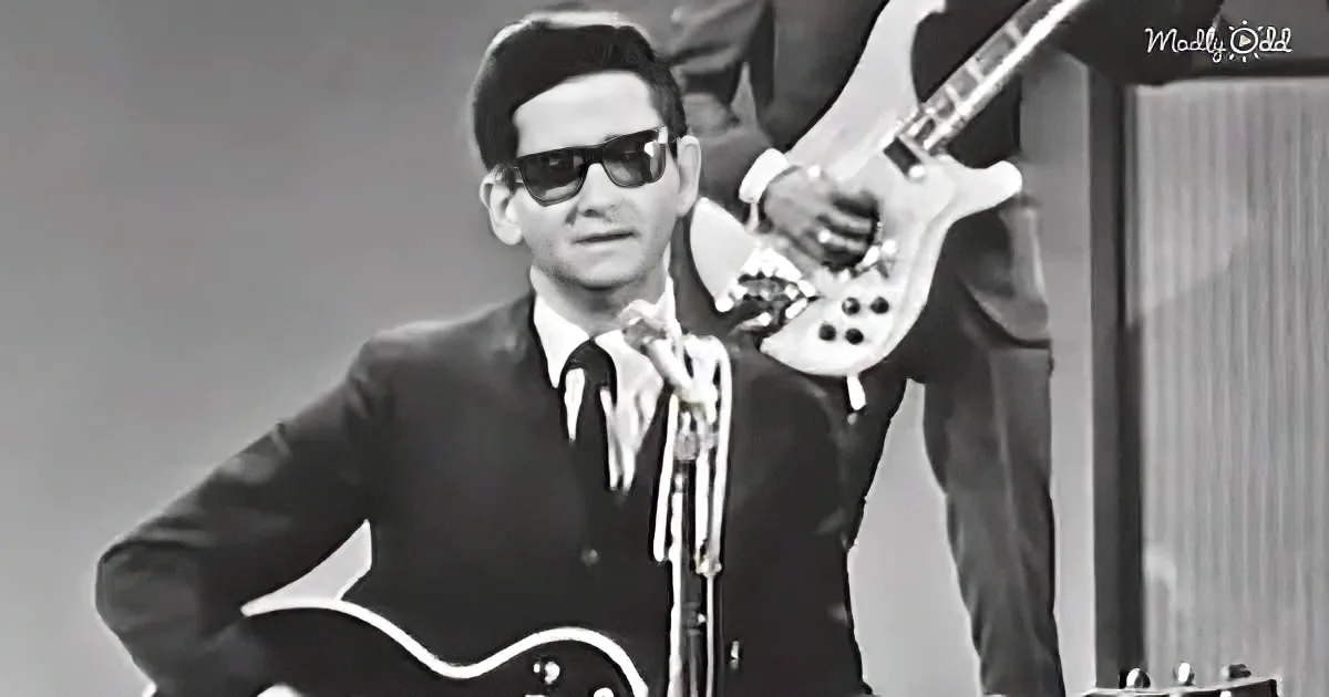 A man in sunglasses and a suit plays a guitar on stage, with another musician in the background.