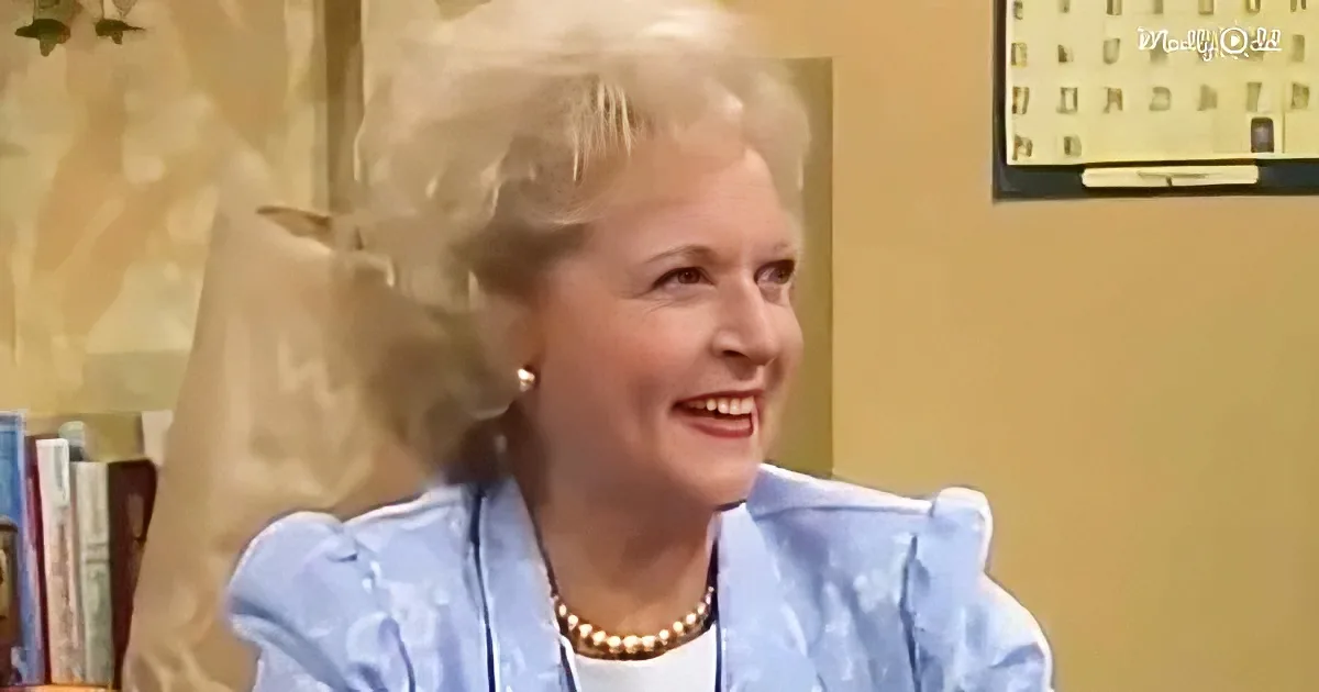 A smiling woman with blonde hair, wearing a blue jacket and pearl necklace, sits in a room with books and a calendar.