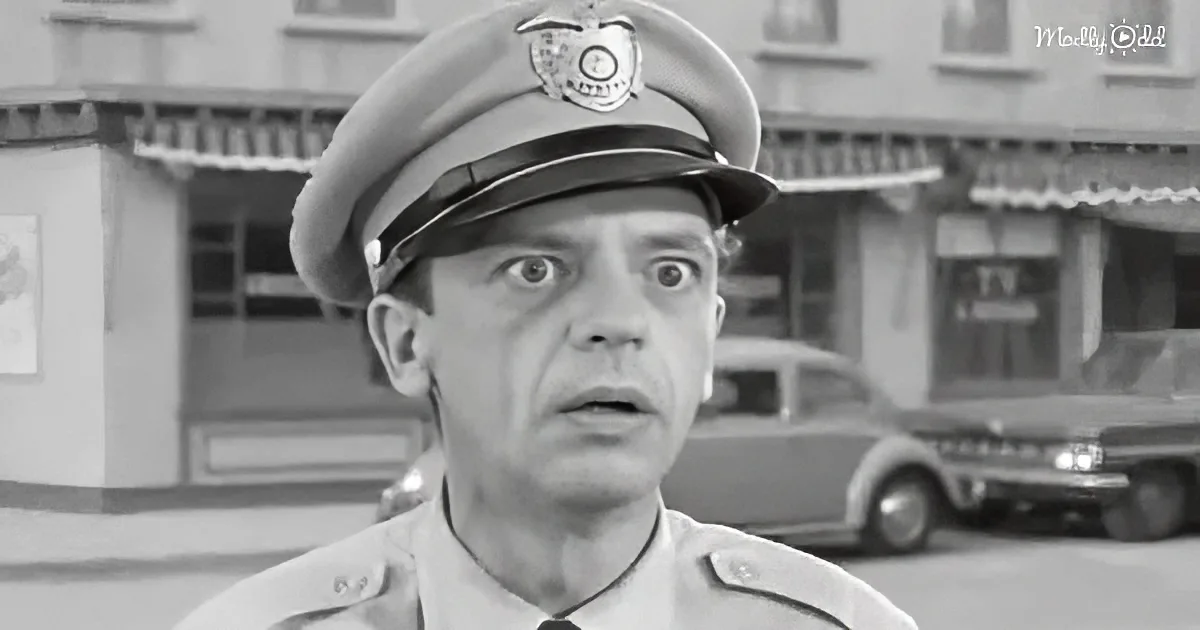 A man in a police uniform looks surprised, standing in front of a building with vintage cars.