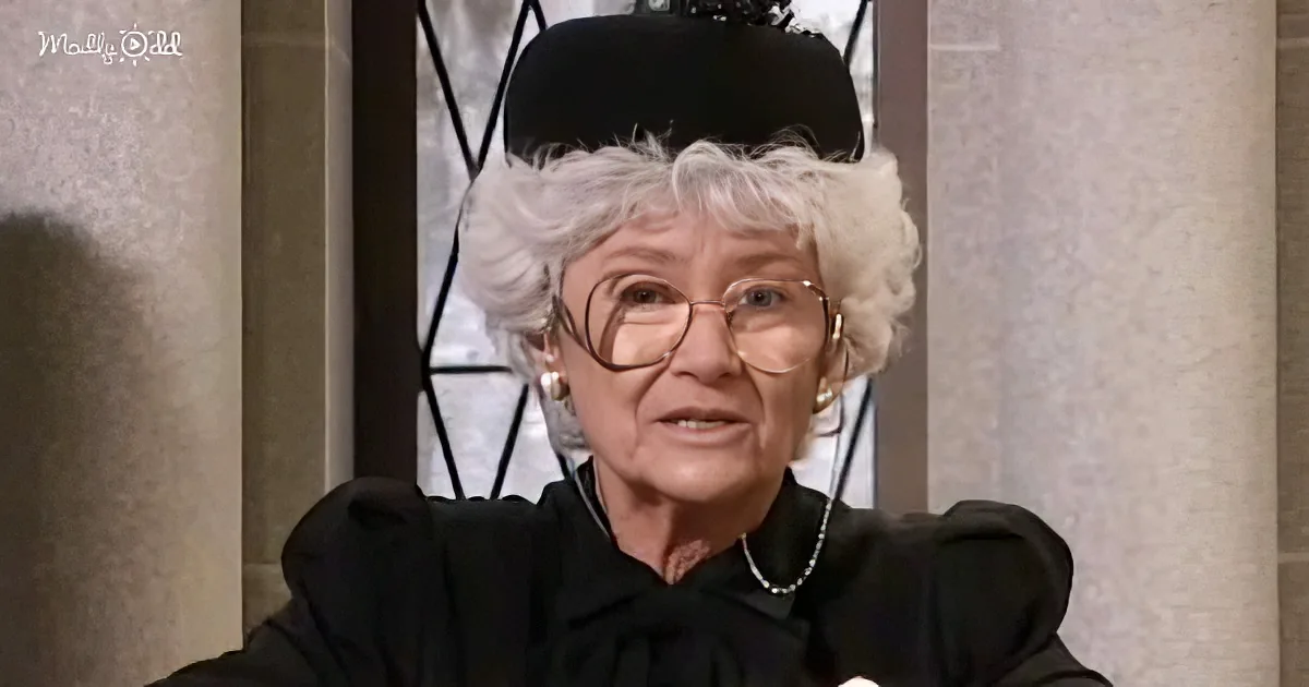 An elderly woman with white hair, glasses, and a black outfit, sitting in front of a window with a decorative frame.