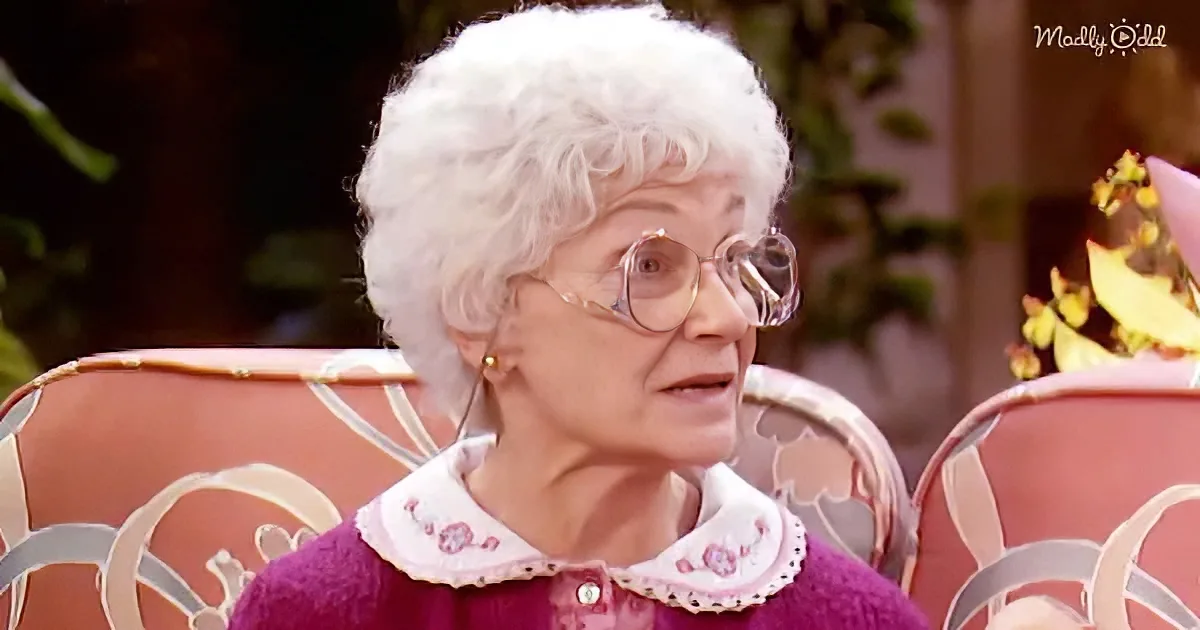 An elderly woman with white hair and glasses, wearing a purple sweater with a white collar, sits in a decorative chair.
